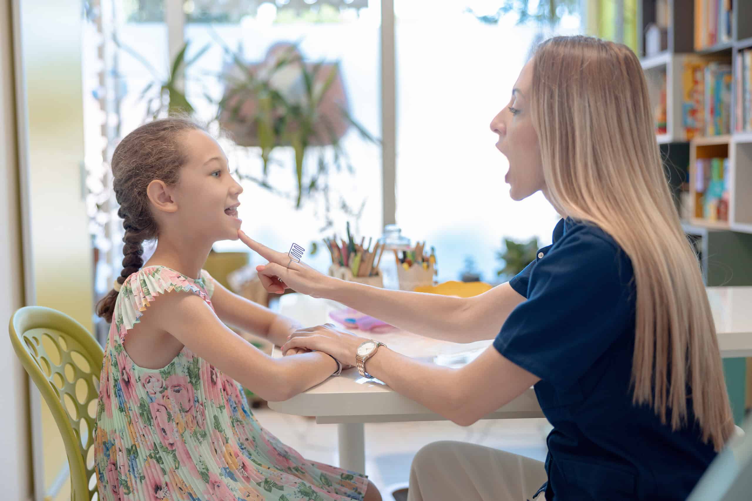 Female speech therapist articulating sounds with elementary age girl and touching her mouth, speech therapy techniques
