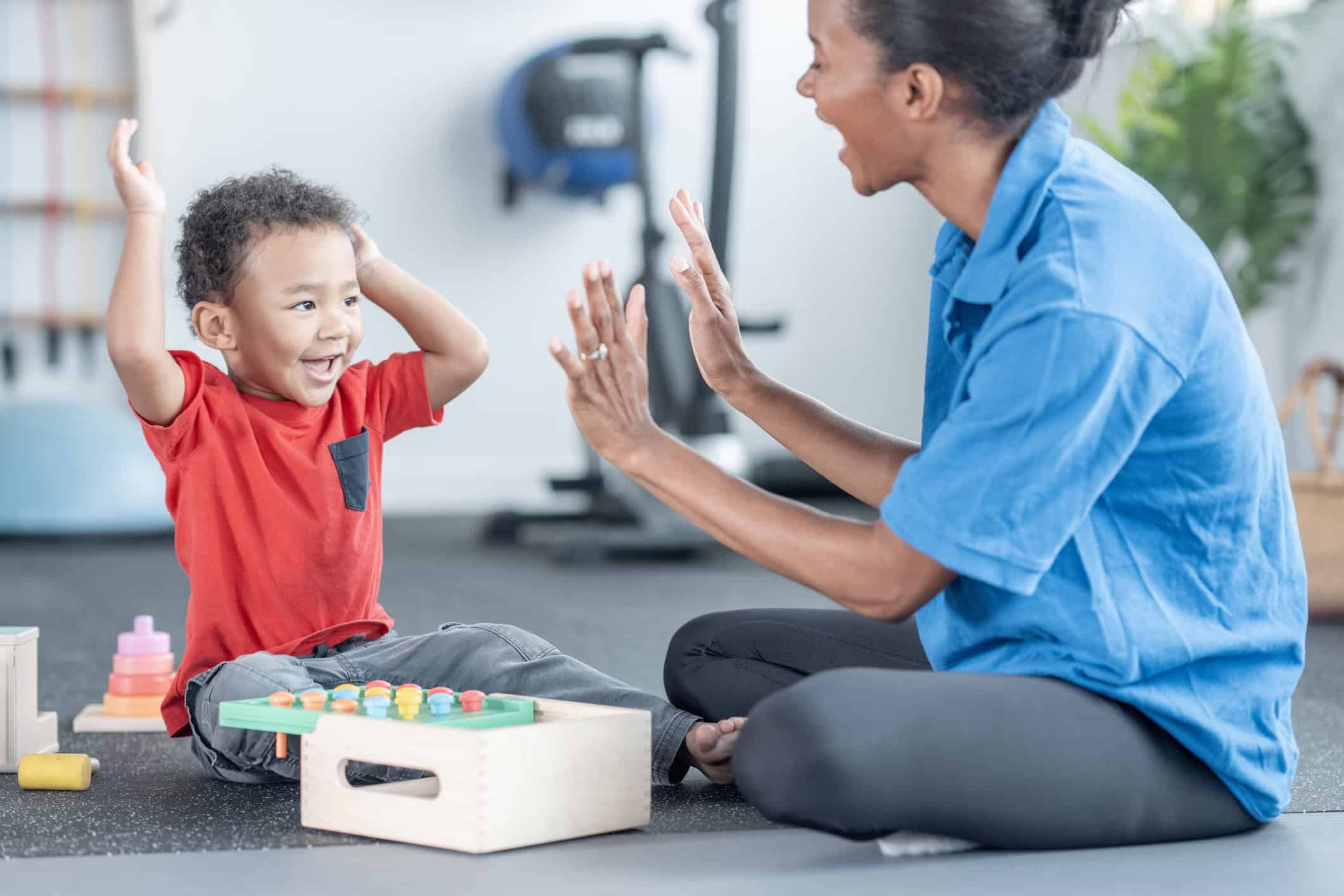 A happy young Black boy in a red shirt and a woman in a blue top celebrate with a high five during an indoor play therapy session, surrounded by educational toys.